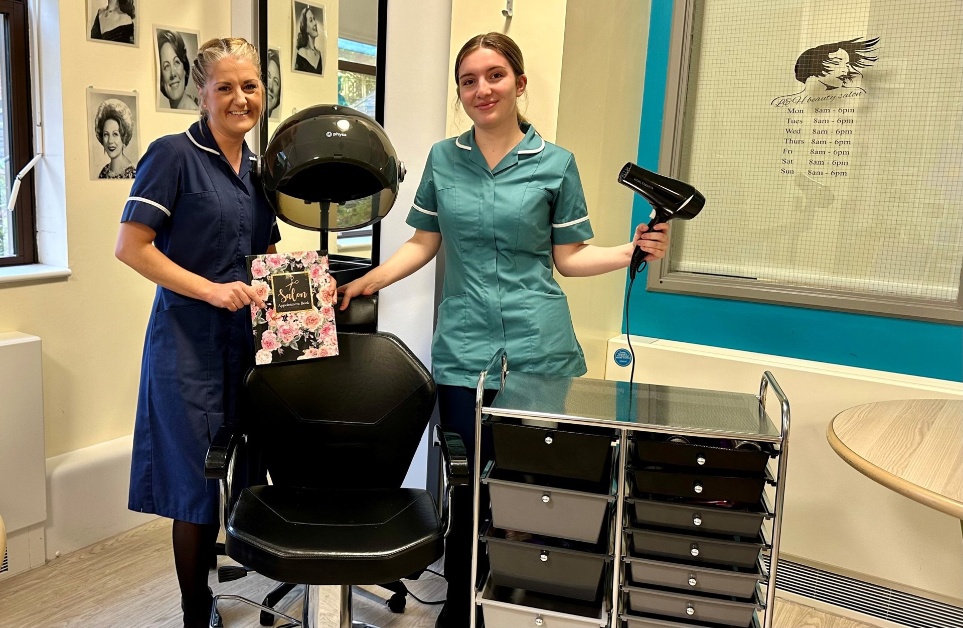 Forget me not ward matron, wearing dark blue uniform and healthcare assistant in turquoise uniform holding a hairdryer. Both stood next to a salon chair at Warrington Hospital's forget me not unit.