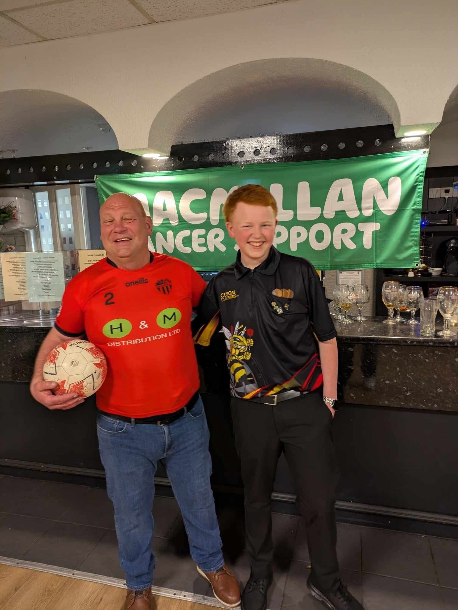 Warren Bibby wearing a red T-shirt with a football in his arms. Warren is stood with a young adult male who is wearing a black drats T-shirt.